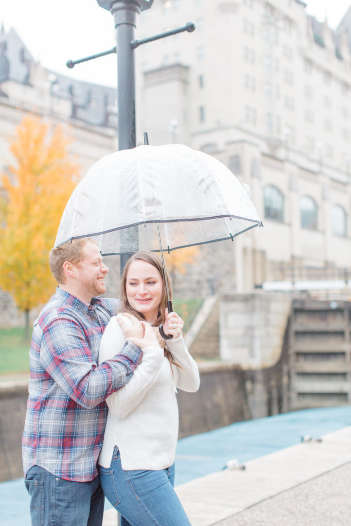 Couple standing in the rain during engagement session with the Locks and Chateau Laurier in the background having fun with each other